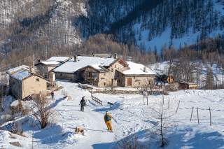 Mehrere Skifahrer auf einem verschneiten Bergweg in der Abfahrt vor einem kleinen Bergdorf mit schneebedeckten Steinhäusern, umgeben von winterlichen, kargen Bäumen.