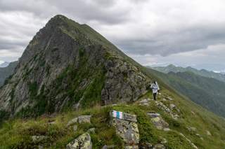 Wanderer mit Trekkingstöcken auf schmalem Bergpfad unter bewölktem Himmel, im Hintergrund steiler, grüner Felsgipfel.