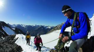 Drei Menschen sind auf Hochtour, sie sind verbunden durch ein Seil und steigen durch den Schnee bergauf.