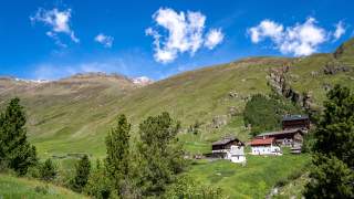 Blick auf sommerlich grüne Hänge. Man sieht ein paar alte Höfe und eine Kapelle.