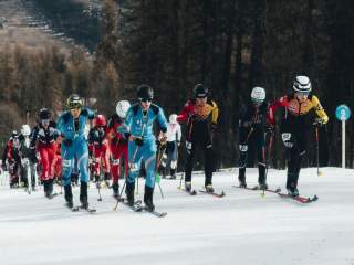 mehrere Skimo Athleten, die nebeneinander und hintereinander die Piste hochgehen. im Hintergrund sieht man einen Wald und Wiesen im Tal. Zwei deutsche Athleten laufen an der Spitze mit. Daneben läuft ein Italiener in einem blauen Rennanzug.