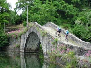 Zwei Mountainbiker fahren über eine gebogene altertümliche Steinbrücke.