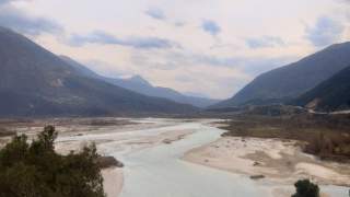 Fluss fließt durch weites, sandiges Flusstal umgeben von hohen Bergen unter bewölktem Himmel.
