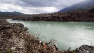 Fluss mit trübem, grünlichem Wasser fließt zwischen felsigen Ufern unter bewölktem Himmel, an den Ufern hängen Plastiksäcke und Müll in den Büschen.