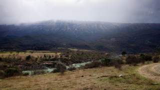 Fluss, der sich durch eine bewaldete Hügellandschaft mit Bergen im Hintergrund windet, unter bewölktem Himmel.