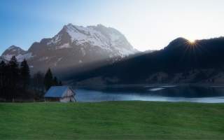 Sonnenaufgang hinter bewaldetem Hügel, schneebedeckte Berge im Hintergrund, ein See und eine kleine Holzhütte auf einer Wiese im Vordergrund.