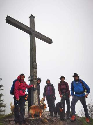 Wandergruppe mit Hunden an einem Gipfelkreuz bei Regenwetter