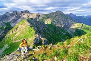 Zwei Wanderer gehen einen steilen Gratweg zu einem Alpengipfel. Die Sonne scheint zwischen den Wolken hindurch