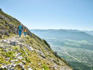 Zwei Wanderer mit Rucksäcken und Wanderstöcken gehen auf einem Pfad am Berghang, im Hintergrund ist eine Stadt in einem Tal mit Bergpanorama zu sehen.