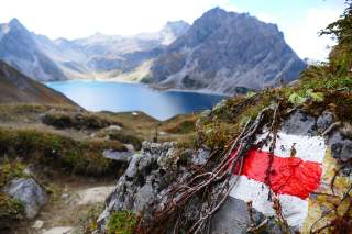 Im Hintergrund ein von Bergen eingerahmter See, im Vordergrund eine rot-weiß-rote Wanderwegmarkierung.