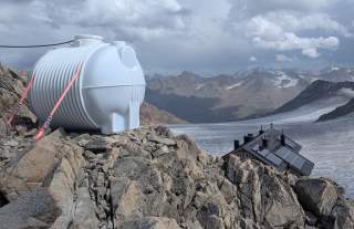 Großer, weißer Kunststofftank auf felsigem Berggipfel, im Hintergrund Berghütte mit Solarpanels und Gletscherlandschaft.