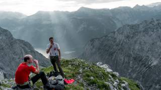 Zwei Wanderer essen eine Kleinigkeit mit Blick auf Königssee und Berge