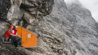 Zwei Bergsteiger machen Pause an orangener Biwakschachtel