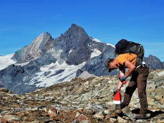 Ein Wanderer mit Rucksack markiert auf einem felsigen Bergpfad einen rot-weiß-rot markierten Stein, im Hintergrund sind schneebedeckte Berggipfel unter blauem Himmel zu sehen.