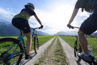 Zwei Personen fahren mit Mountainbikes auf einem Schotterweg durch eine grüne Wiese, im Hintergrund Berge und blauer Himmel.