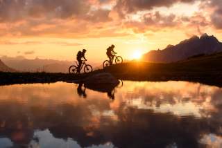 Zwei Menschen auf Mountainbikes vor Bergsee im Sonnenuntergang