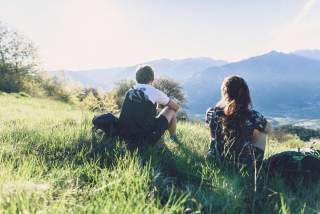 Ein junger Mann und eine junge Frauen sitzen, mit dem Rücken zum Betrachter, auf einer Bergwiese. Sie wirken gedankenversunken in die Berge schauend.