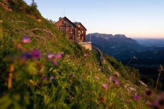 Hütte auf einem grünen Berghang mit bunten Wildblumen im Vordergrund und einer Bergkette im Hintergrund bei Sonnenuntergang.