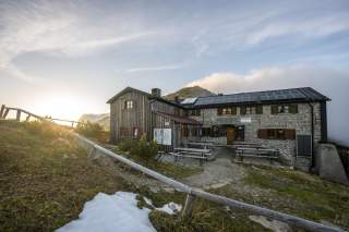 Berghütte aus Stein und Holz, umgeben von einer Berglandschaft bei Sonnenuntergang.
