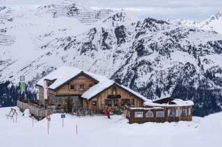 Verschneite Berghütte mit Terrasse und Personen in Skikleidung vor schneebedeckten Alpen im Hintergrund.