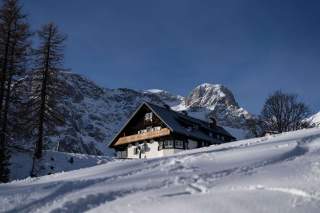 Schneebedeckte Hütte vor schroffen, verschneiten Berggipfeln unter klarem, blauem Himmel. Vereinzelt stehende kahle Bäume umgeben das Haus.