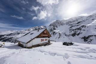 Schneebedecktes Berghaus mit roter Fensterläden vor schneebedeckten Bergen unter bewölktem Himmel.