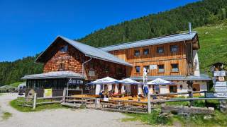 Berghütte mit Sonnenschirmen vor einem bewaldeten Hang an einem sonnigen Tag.