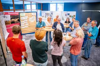 Eine Gruppe von Menschen steht in einem Raum um Tische und Pinnwände. Die Umgebung wirkt wie eine Workshop- oder Besprechungssituation.