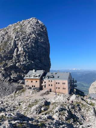 Berghütte mit Holzfassade steht vor einer steilen Felswand, umgeben von felsiger Landschaft und blauem Himmel.
