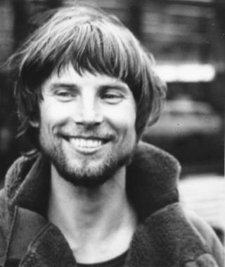 Schwarz-weiß-Porträt eines jungen Mannes mit kurzem Haar und Bart, der breit lächelnd nach links schaut. Er trägt einen warmen Mantel mit hochgestelltem Kragen.