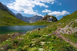 Sommerliche Alpenlandschaft mit einem See, an dem eine Alpenvereinshütte steht, dahinter steile Felsflanken.