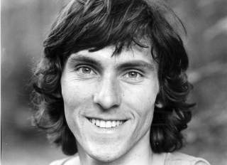 Schwarz-weiß-Porträt eines lächelnden Mannes mit mittellangen, gewellten Haaren vor unscharfem Hintergrund.