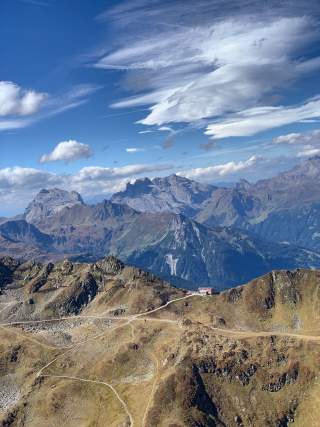 Landschaftsaufnahme von Bergen mit Wanderwegen im Vordergrund und imposanten Gipfeln im Hintergrund unter einem blauen Himmel mit Wolken. Auf einem Hochplateau steht ein kleines Berghaus.