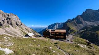 Berghütte mit Solarpanels auf dem Dach in einer grünen Berglandschaft, umgeben von steilen Felswänden unter klarem blauem Himmel.