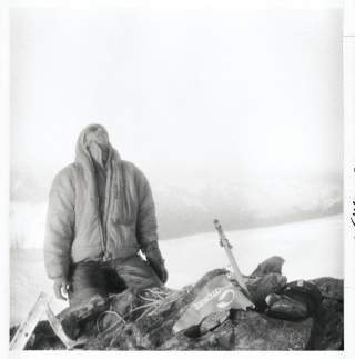 Schwarz-Weiß-Foto eines Bergsportlers in dicker Winterjacke und Hose, die auf einem felsigen Gipfel steht und nach oben blickt; neben der Person liegen Seil und Ski.