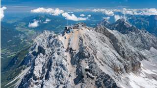 Luftaufnahme eines schroffen, felsigen Berggipfels mit Gebäuden auf dem höchsten Punkt, im Hintergrund grüne Täler und weitere Berge unter blauem Himmel mit wenigen Wolken.