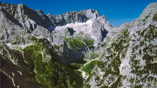Hohe, felsige Berge mit grünen Hängen und einem schmalen, gewundenen Tal im Vordergrund unter blauem Himmel.