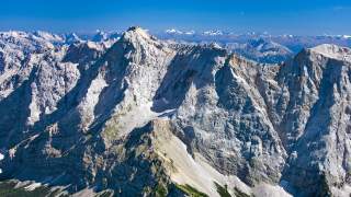 Weite, felsige Alpenlandschaft mit schroffen, grauen Berggipfeln unter blauem Himmel.