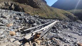 Eine beschädigte Holzbrücke liegt schief über ein felsiges Flussbett in einer bergigen Landschaft.