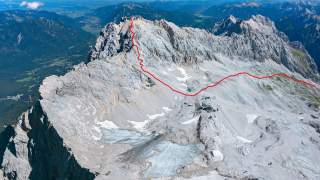 Blick von oben auf ein felsiges Hochgebirge mit Gletscherflächen; eine rote Linie markiert einen Wander- oder Kletterweg über den Grat.