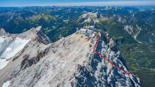Luftaufnahme eines felsigen Bergkamms mit Gebäuden auf dem Gipfel, im Hintergrund bewaldete Berge; ein roter Pfad ist auf den Felsen markiert.