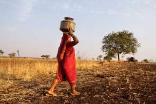 Eine Frau in rotem, traditionellem Kleid trägt ein kleines Wasserfass durch die ausgedorrte Landschaft.