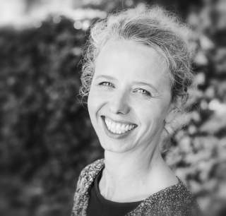 Porträt einer lächelnden Frau mit hellem, lockigem Haar vor verschwommenem Hintergrund.