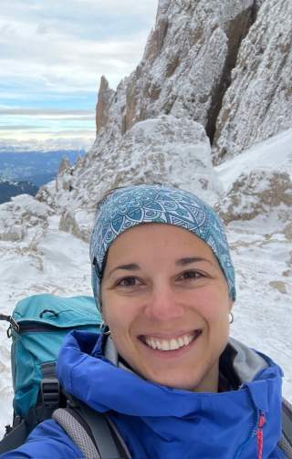 Frau mit blauem Stirnband und blauer Outdoor-Jacke macht Selfie vor verschneiten Felsen in den Bergen.