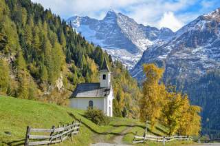 Eine kleine Kapelle steht vor einer schroffen Bergkulisse. Die Berge sind mit Schnee bedeckt, die Bäume färben sich schon herbstlich golden.