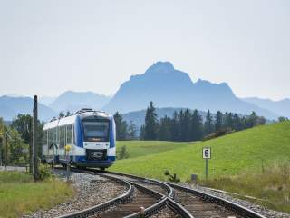 Ein (blau-weißer) Zug fährt durch das hügelige Allgäu. Im Hintergrund hohe Berge.