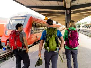 Oftmals geht's per Bahn schneller und bequemer in die Berge. Drei Wanderer laufen auf Bahnsteig