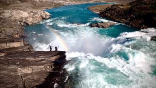 Zwei Personen mit Kajakausrüstung stehen am Rand eines tosenden Wasserfalls, neben ihnen erscheint ein Regenbogen im Sprühnebel. Der Fluss umgibt felsige Ufer in einer weiten, kargen Landschaft.