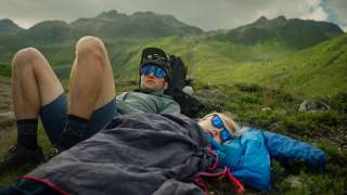 Zwei Personen liegen entspannt in einer Berglandschaft, eine ist in einem Schlafsack eingehüllt, beide tragen Sonnenbrillen. Im Hintergrund sind grüne Berge unter bewölktem Himmel zu sehen.