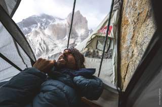 Ein Mann liegt in einer Hängematte  an einer Felswand, trägt eine Daunenjacke und blickt nach oben, im Hintergrund sind schneebedeckte Berge zu sehen. Neben ihm ist ein Seil und eine an der Wand befestigte Hülle.